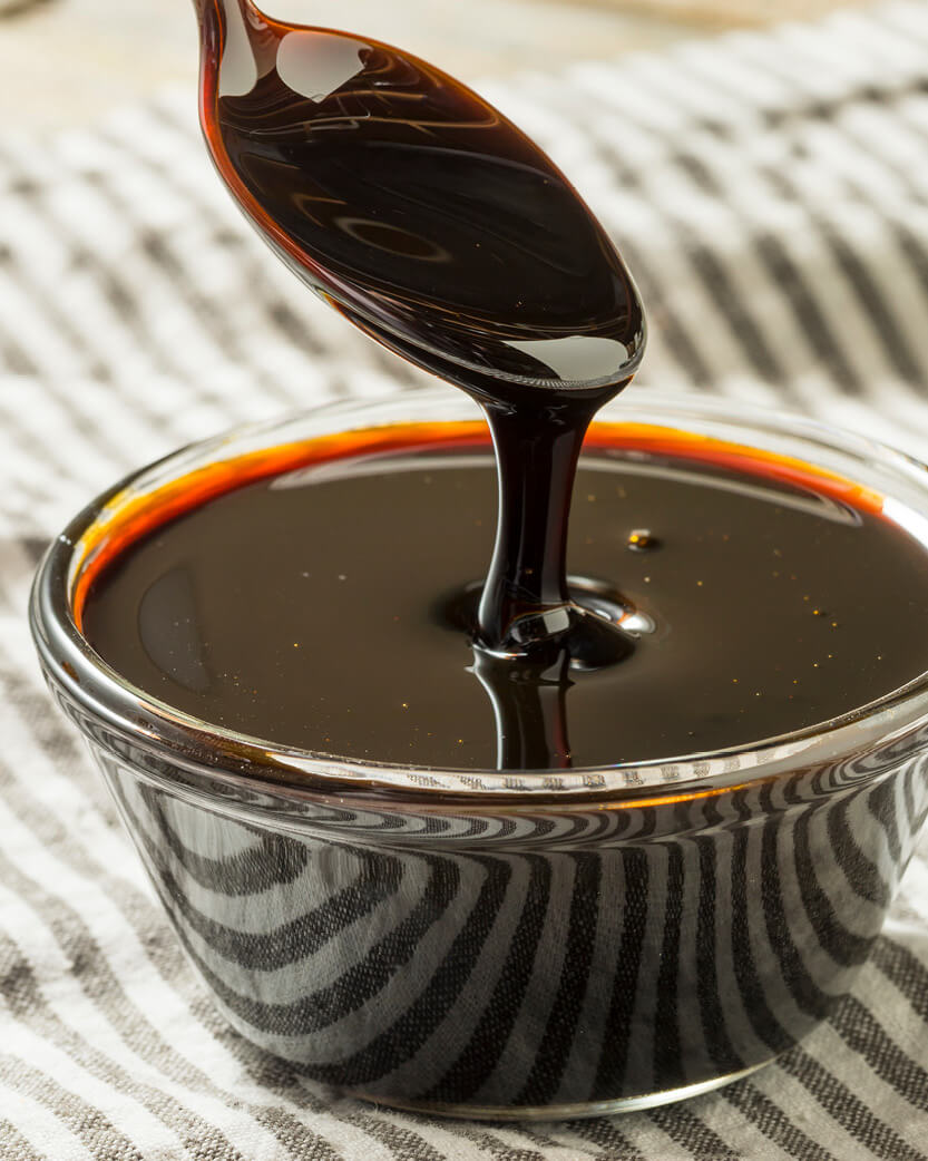 Dark syrup being poured from a spoon into a glass bowl on a striped fabric background