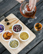 bowls of dried herbs with a glass pot of tea being poured into a cup on a dark wooden table. 