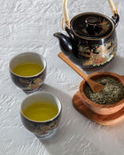 Tea set with two cups, a teapot, and a bowl of tea leaves on a textured white surface.