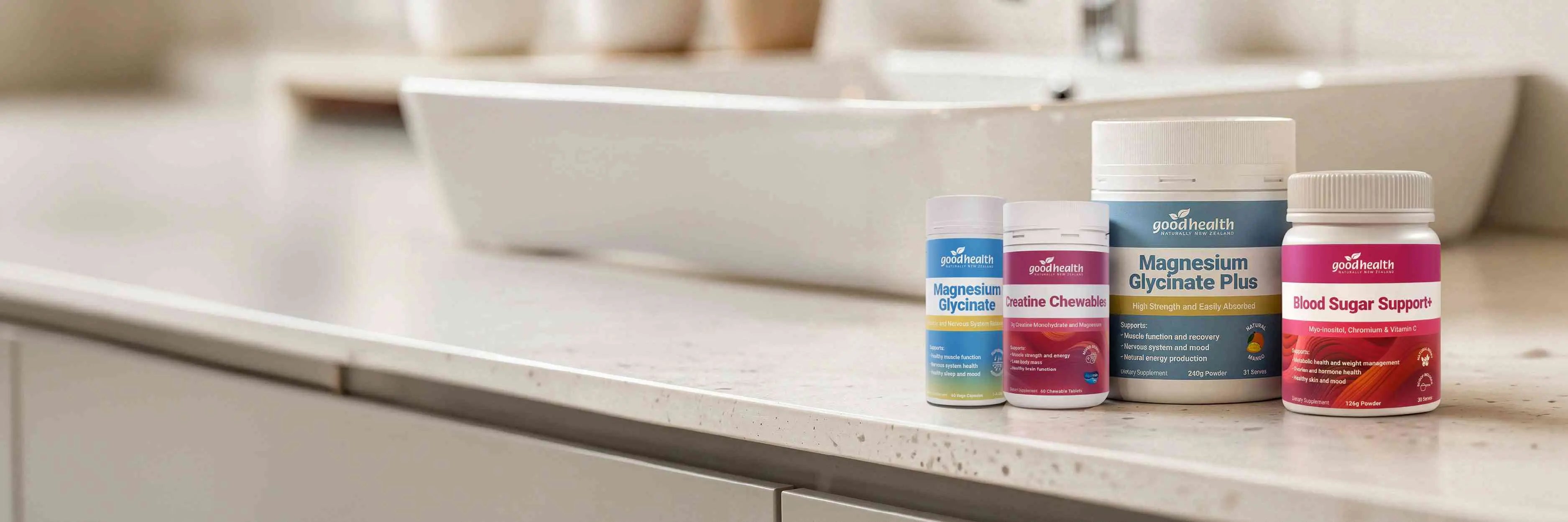 Four supplement bottles from Good Health arranged on a bathroom countertop in front of a sink. The products include Magnesium Glycinate, Magnesium Glycinate Plus, Blood Sugar Support and Creatine Chewables.