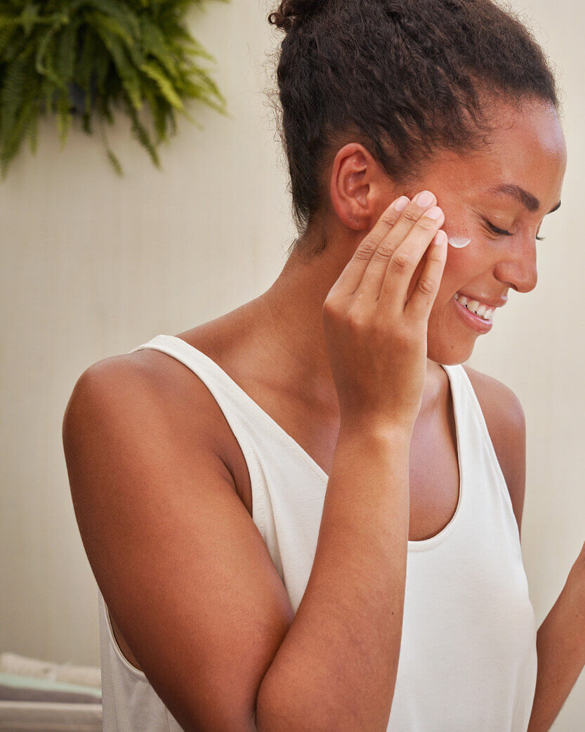 Woman applying cream to her face with a blurred plant in the background.