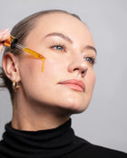 Woman applying a dropper of liquid to her face against a neutral background.
