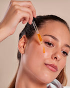 Person applying a dropper of liquid to their face against a neutral background.