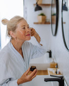 Woman in a bathrobe applying cream to her face in front of a mirror.