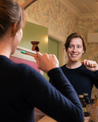 Woman brushing her teeth in front of a mirror with floral wallpaper.