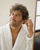 Man in a white robe cleaning his ear with a small mirror.