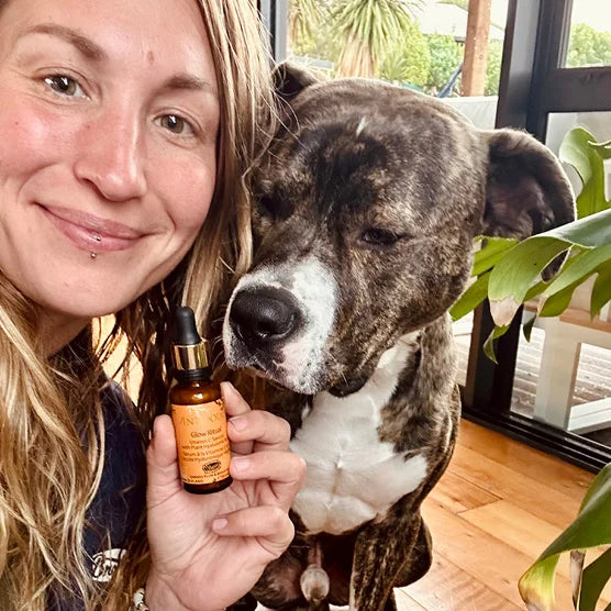 A woman holds a small amber dropper bottle of Antipodes Glow Vitamin C Serum next to a brindle and white pitbull-type dog in an indoor setting with a plant and window visible.