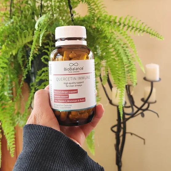 A hand holds a bottle of BioBalance New Zealand Quercetin Immune dietary supplement capsules in front of a green fern plant and a candle holder.