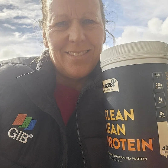 A smiling woman outdoors holds a large tub of GIB Clean Lean Protein powder next to her. She is wearing a black GIB puffer jacket with the GIB logo visible on the chest, and the background shows a cloudy blue sky.