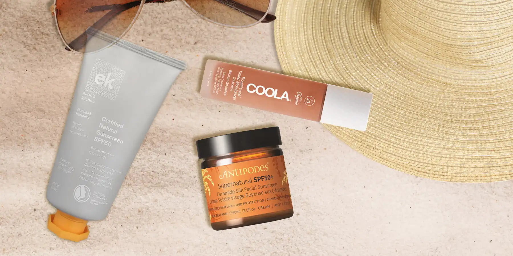 Sunscreen products including a jar, tube, and bottle on a sandy surface with a hat and sunglasses.