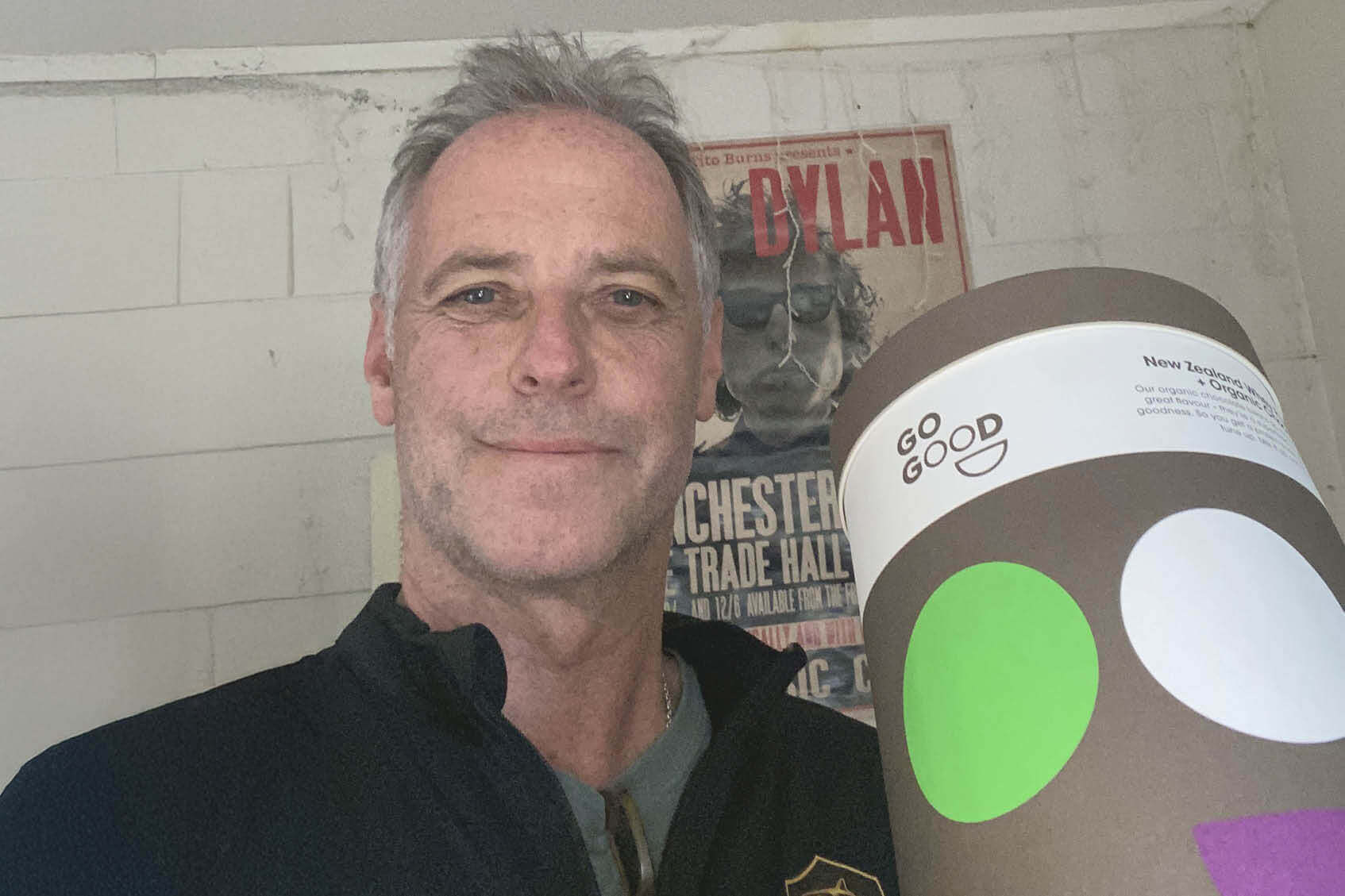 Man holding a cylindrical product with a visible brand logo, standing in front of a wall with posters.