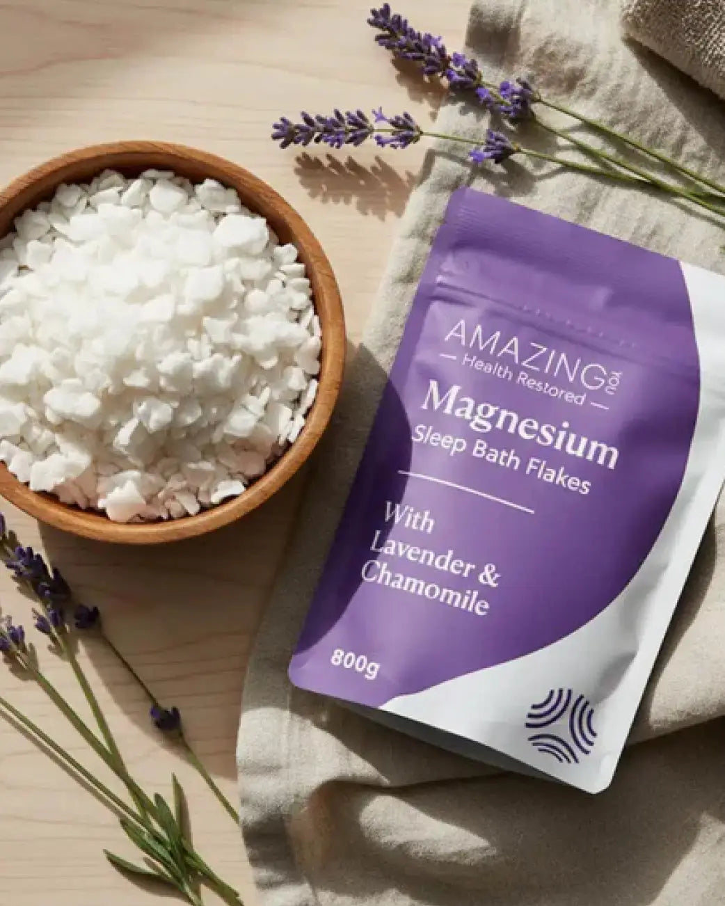 A stand-up pouch of Sleep Bath Flakes with lavender and chamomile, placed on a beige cloth next to a wooden bowl filled with white bath flakes, with lavender sprigs on a light wood surface.