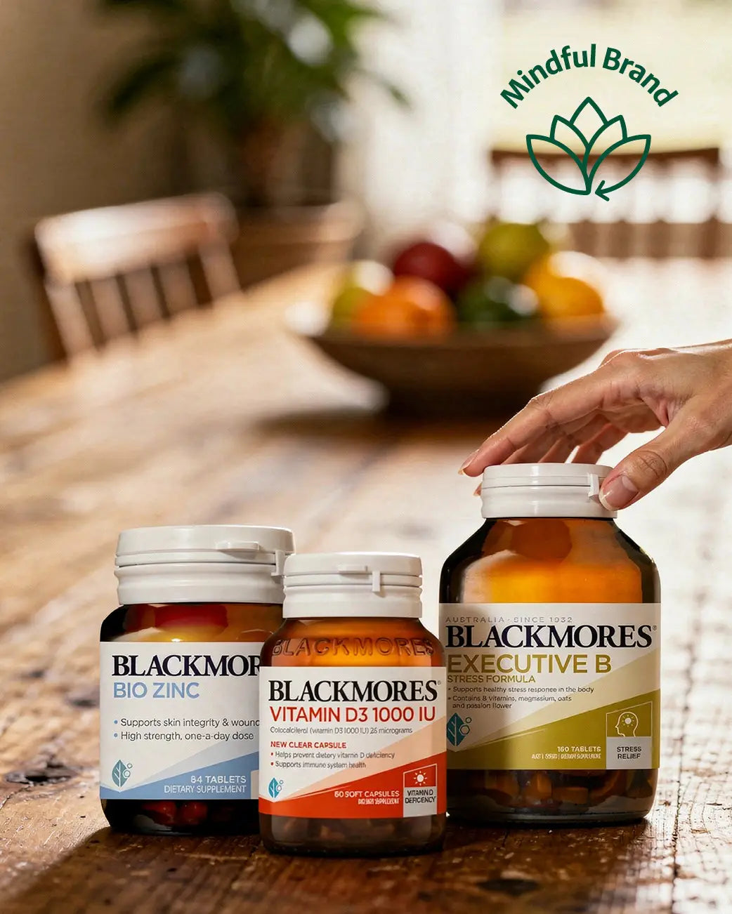 Three Blackmore's vitamin bottles on a wooden table with a blurred background of fruits and chairs.