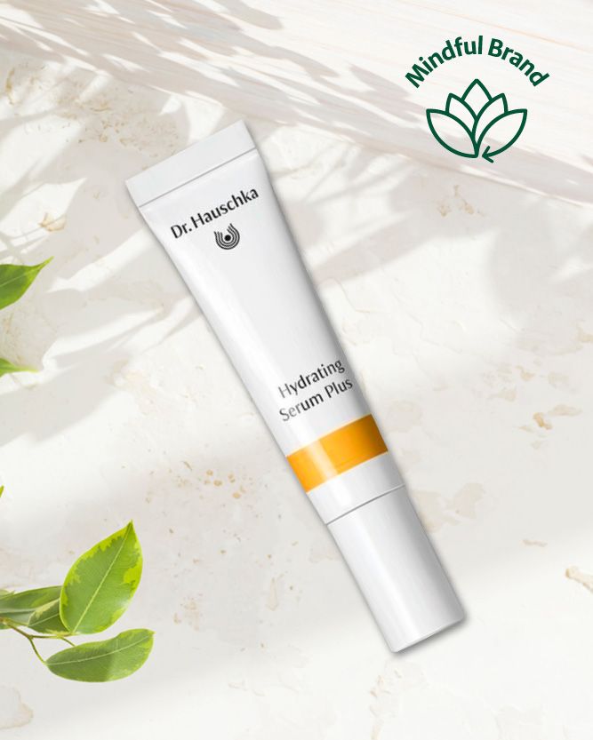 A white tube of Dr. Hauschka Hydrating Serum Plus with a yellow-orange band, styled diagonally on a textured white surface with soft leaf shadows and green leaves in the corners.