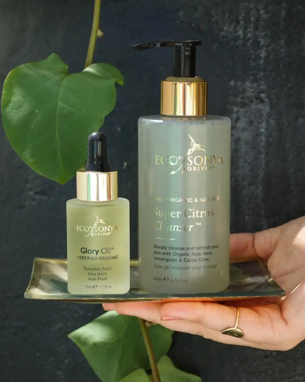 Two skincare products, 'Glory Oil' and 'Super Citrus Cleanser', held by a hand with a dark background.