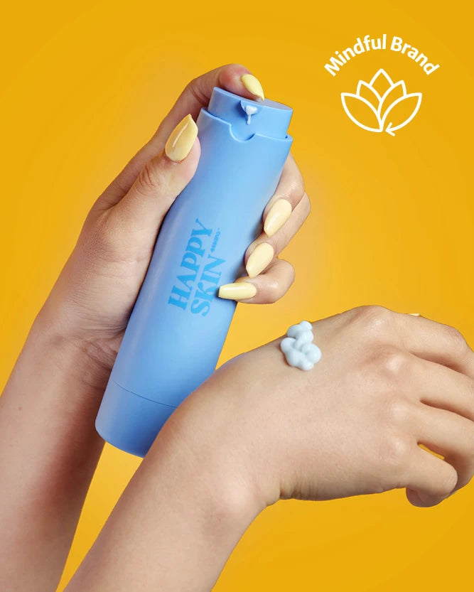 Hand holding essano Happy Skin in a blue pump bottle with some on her other hand. On a yellow background with a mindful brand logo.