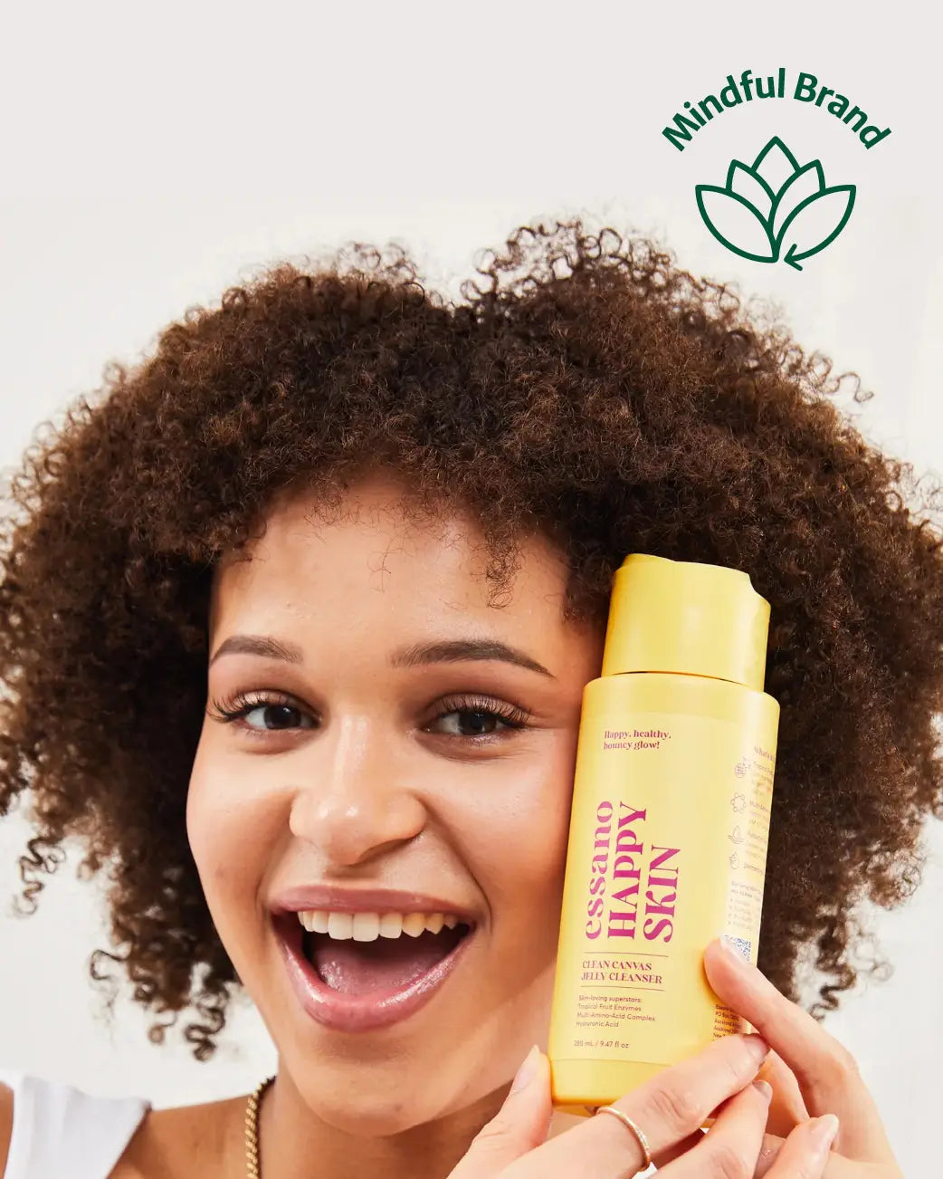 A smiling woman with curly brown hair holds a yellow bottle of cleanser next to her face against a light gray background. A green 'Mindful Brand' logo is visible in the top right corner.