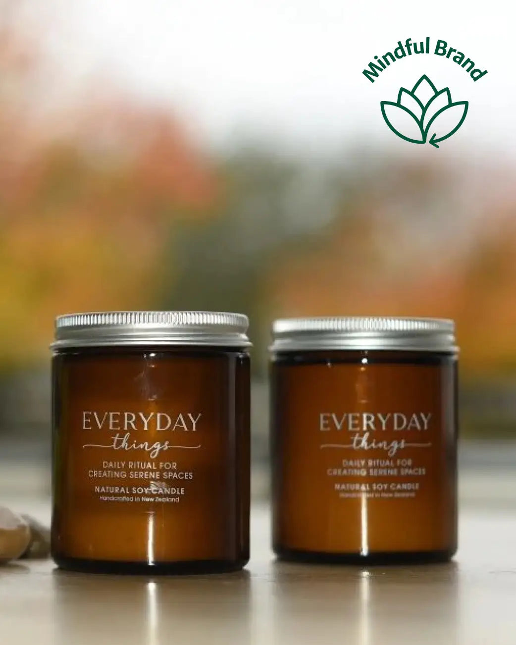 Two amber glass jar candles with silver lids labeled 'EVERYDAY things' sit on a tabletop with a blurred outdoor background. A green 'Mindful Brand' logo appears in the top right corner.