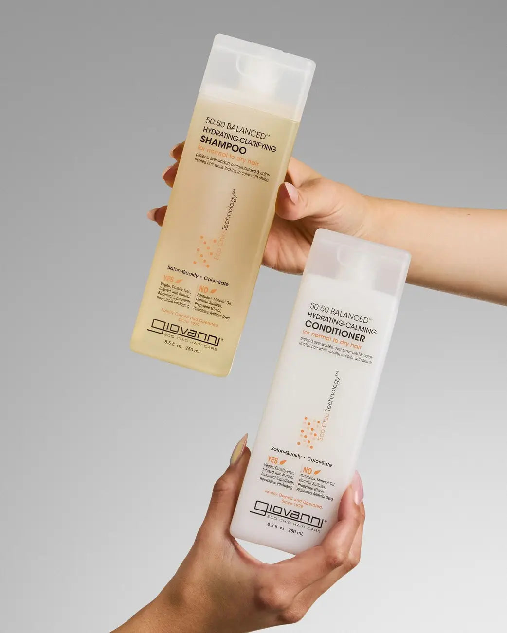 Two hands holding Giovanni 50:50 Balanced Hydrating Shampoo and Conditioner bottles against a neutral gray gradient background.