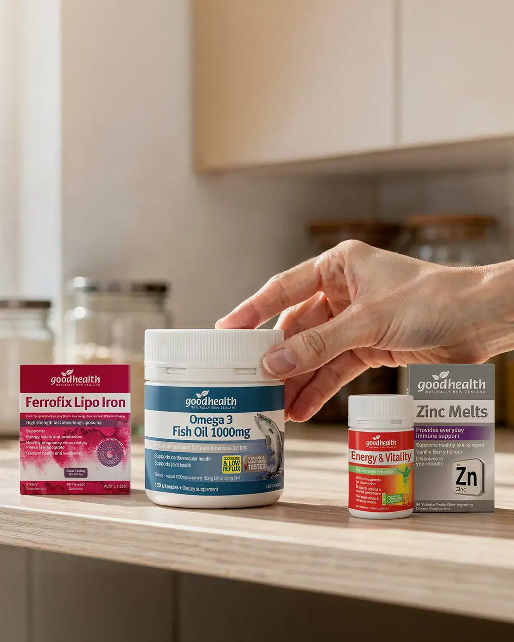 Hand reaching for a container of goodhealth Omega 3 Fish Oil 1000mg on a kitchen counter with other health products.