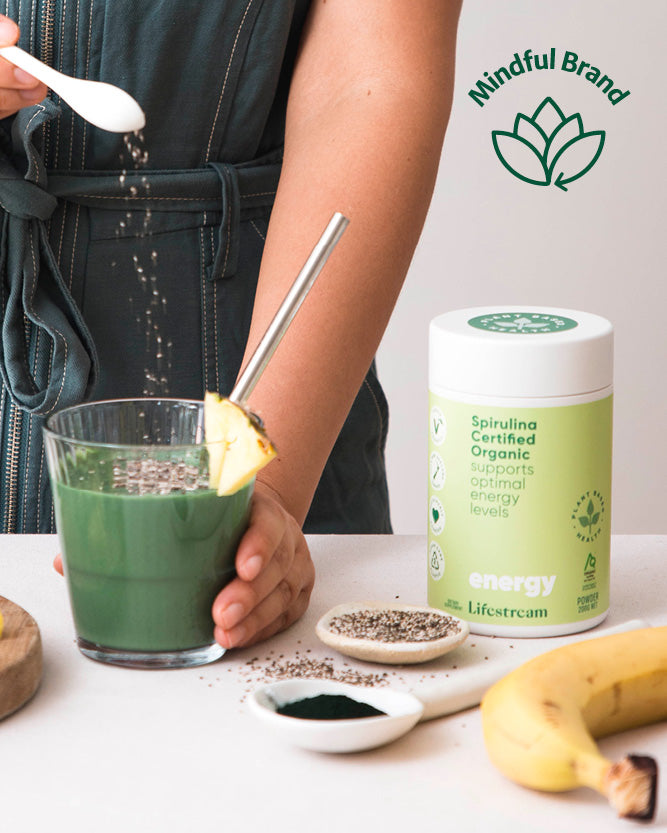 Person holding a green smoothie with a Mindful Brand container and ingredients on a table.