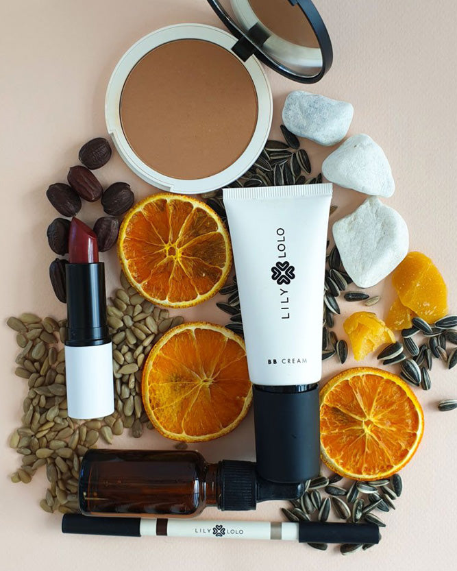 Makeup products including a compact mirror, cream tube, lipstick, and small bottle on a pink background with dried oranges and sunflower seeds.