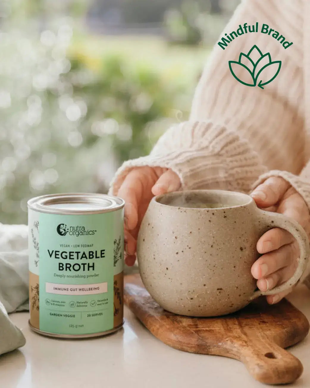 A person in a cream knit sweater holds a speckled beige ceramic mug on a wooden board next to a can of Veggie's Organic Vegetable Broth. The background is a soft, blurred outdoor green scene with a 'Mindful Brand' logo overlay in the top right.