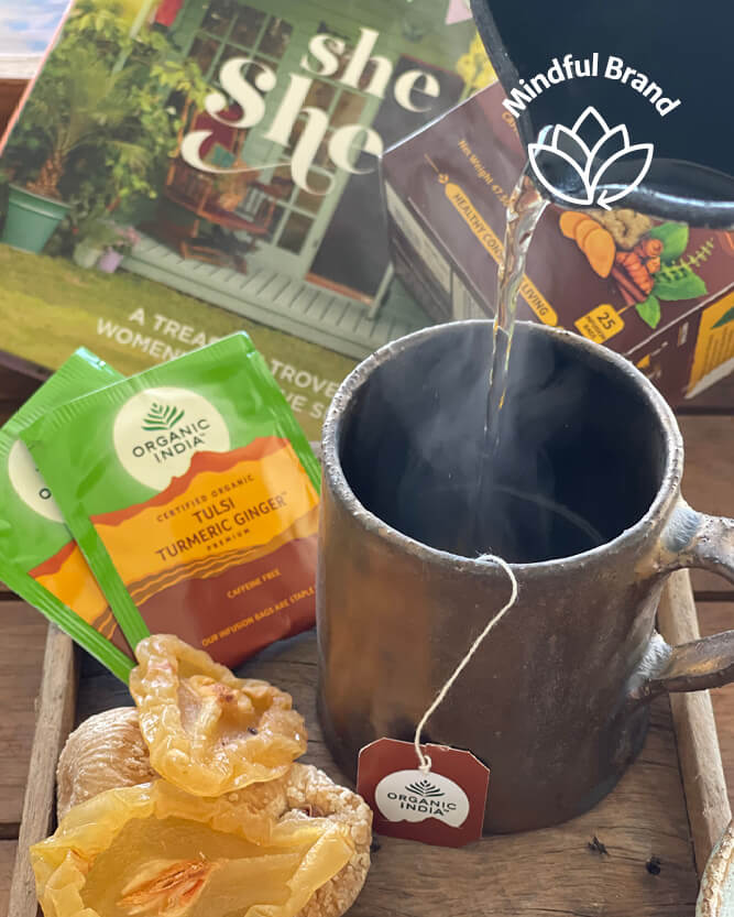 Organic India Tulsi Turmeric Ginger tea bags are shown being brewed into a rustic dark metal mug on a wooden tray, with dried tulsi leaves and a box of the tea in the background.