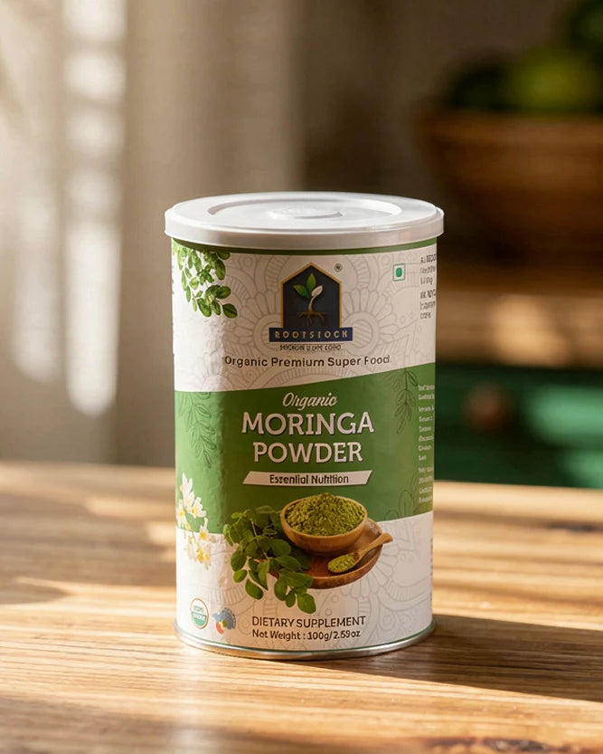 A 130g canister of Rootstock Organic Premium Super Food Moringa Powder sits on a wooden table in warm, natural light. The label is green and white with illustrations of moringa leaves and flowers, and a photo of the green powder in a wooden bowl.