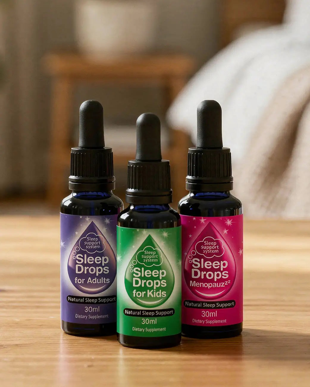 Three small glass dropper bottles of sleep drops arranged on a wooden surface, with a softly blurred indoor background. The bottles have black caps and dark blue glass, and the labels are purple, green, and pink.