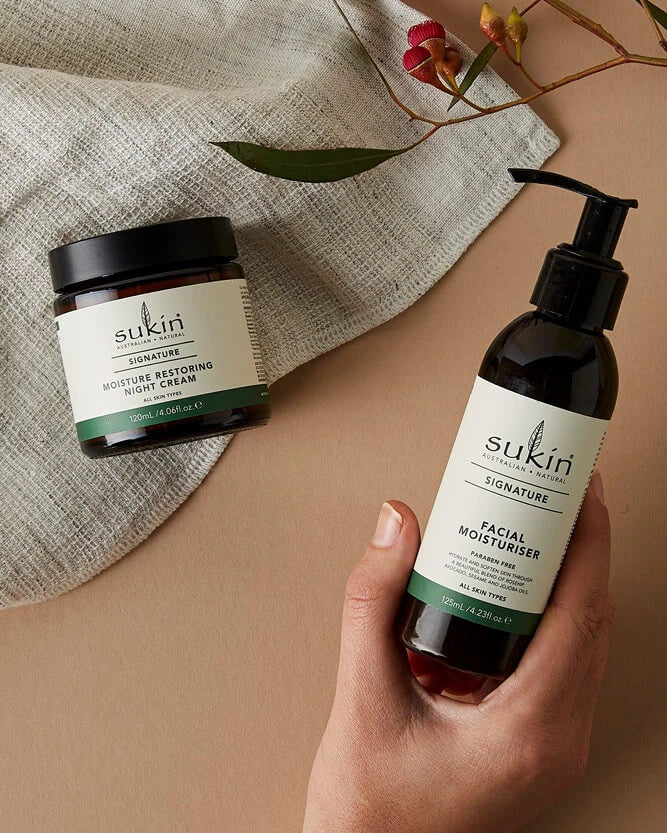 A hand holds a dark amber bottle of Sukin Signature Facial Moisturiser against a beige background, with a second jar of Sukin Moisture restoring night cream resting on a textured linen cloth.