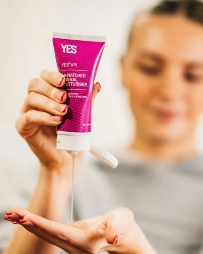 A person holds a squeeze tube of YES! pH Matched Vaginal Moisturiser, with the white cap open and a thin stream of moisturiser dripping onto their palm. The background is softly blurred, showing a woman's face and upper body.