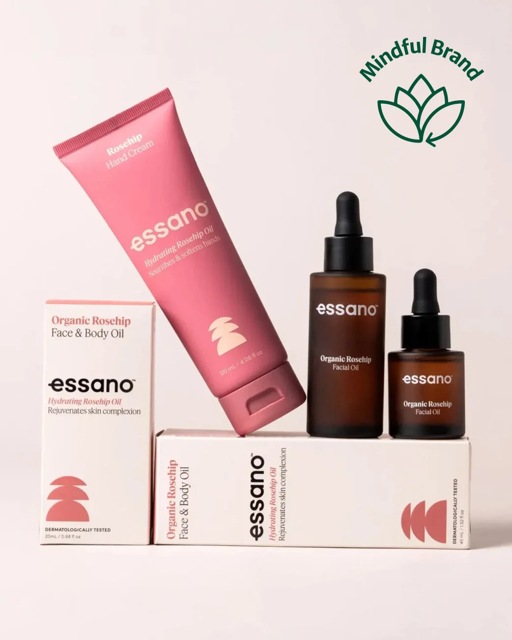 Essano Organic Rosehip Face & Body Oil set including a pink face cream tube, two amber dropper bottles, and white boxes, arranged on a light beige background with a green 'Mindful Brand' logo in the top right.