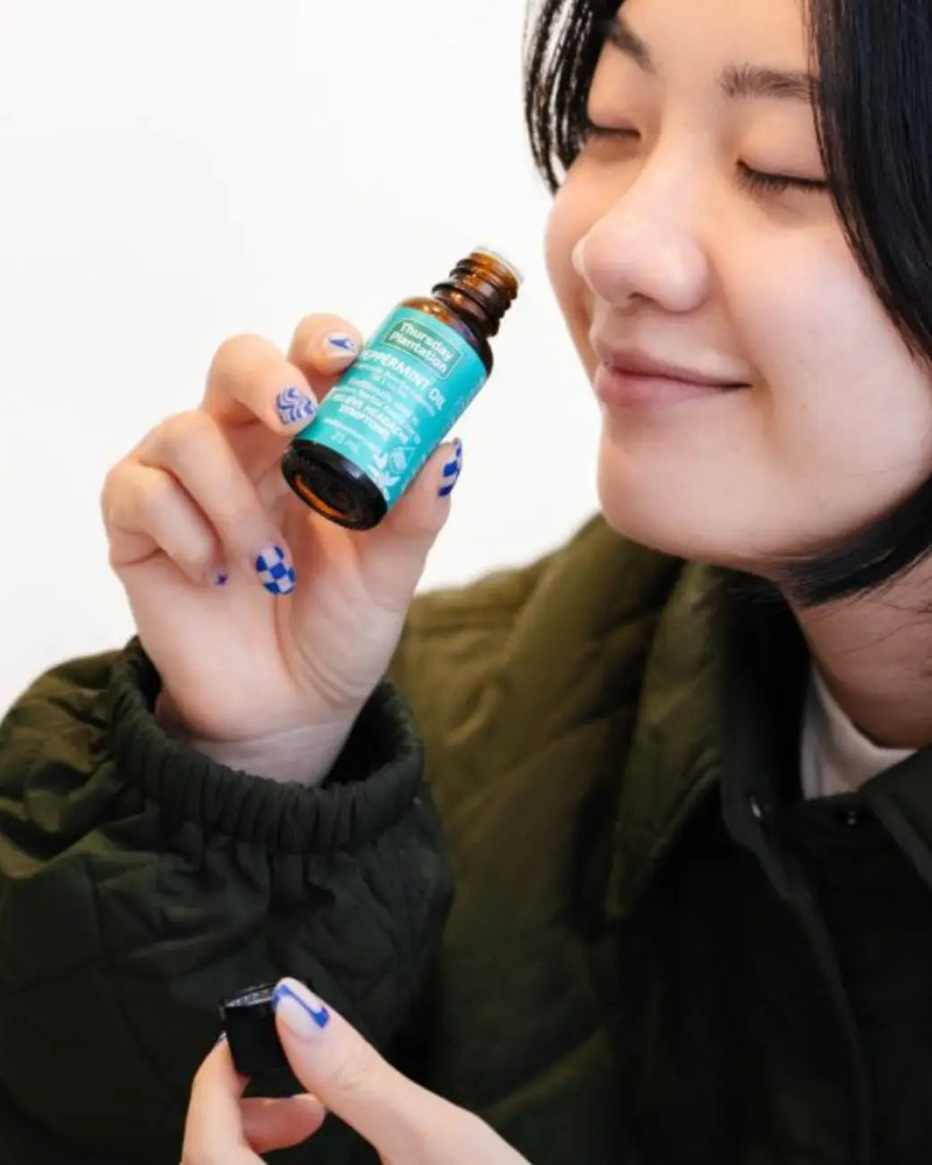 A woman holding a small amber glass bottle with a bright teal label, shown in a close-up lifestyle shot against a plain white background.