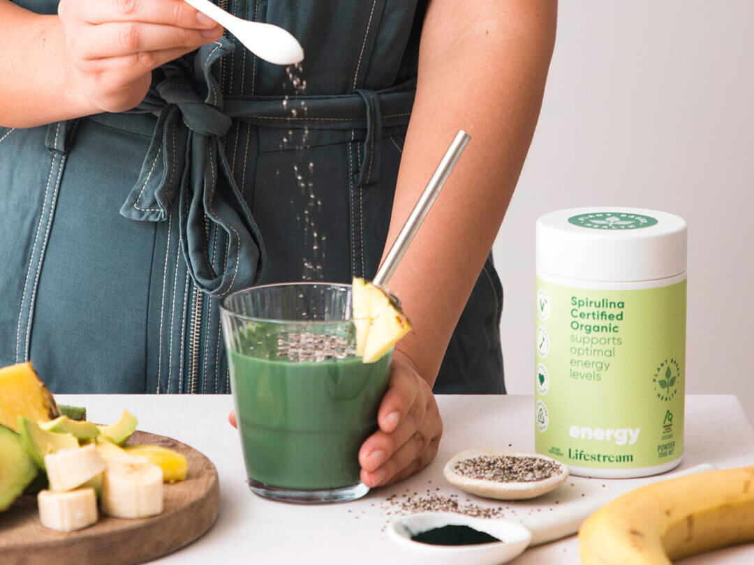 Person making spirulina on a white counter with fresh fruit and vegetables.