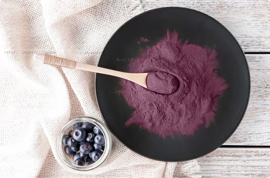 Purple powder on a black plate with a wooden spoon and blueberries on a light wooden surface.