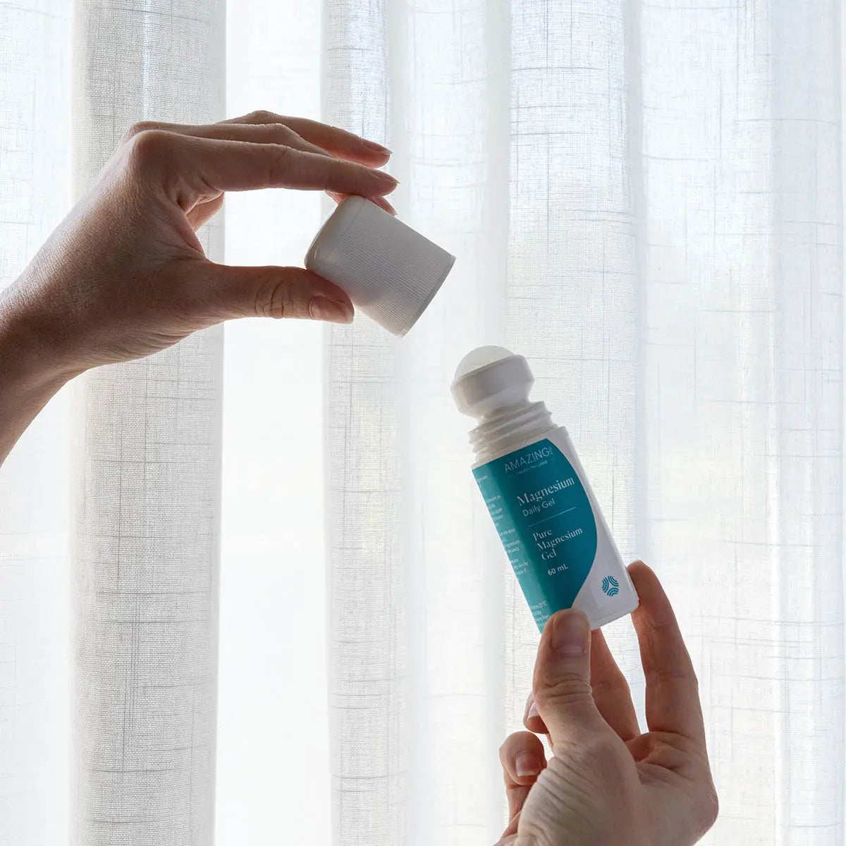 Two hands holding a small bottle of roll on magnesium gel with a blue label against a white curtain background