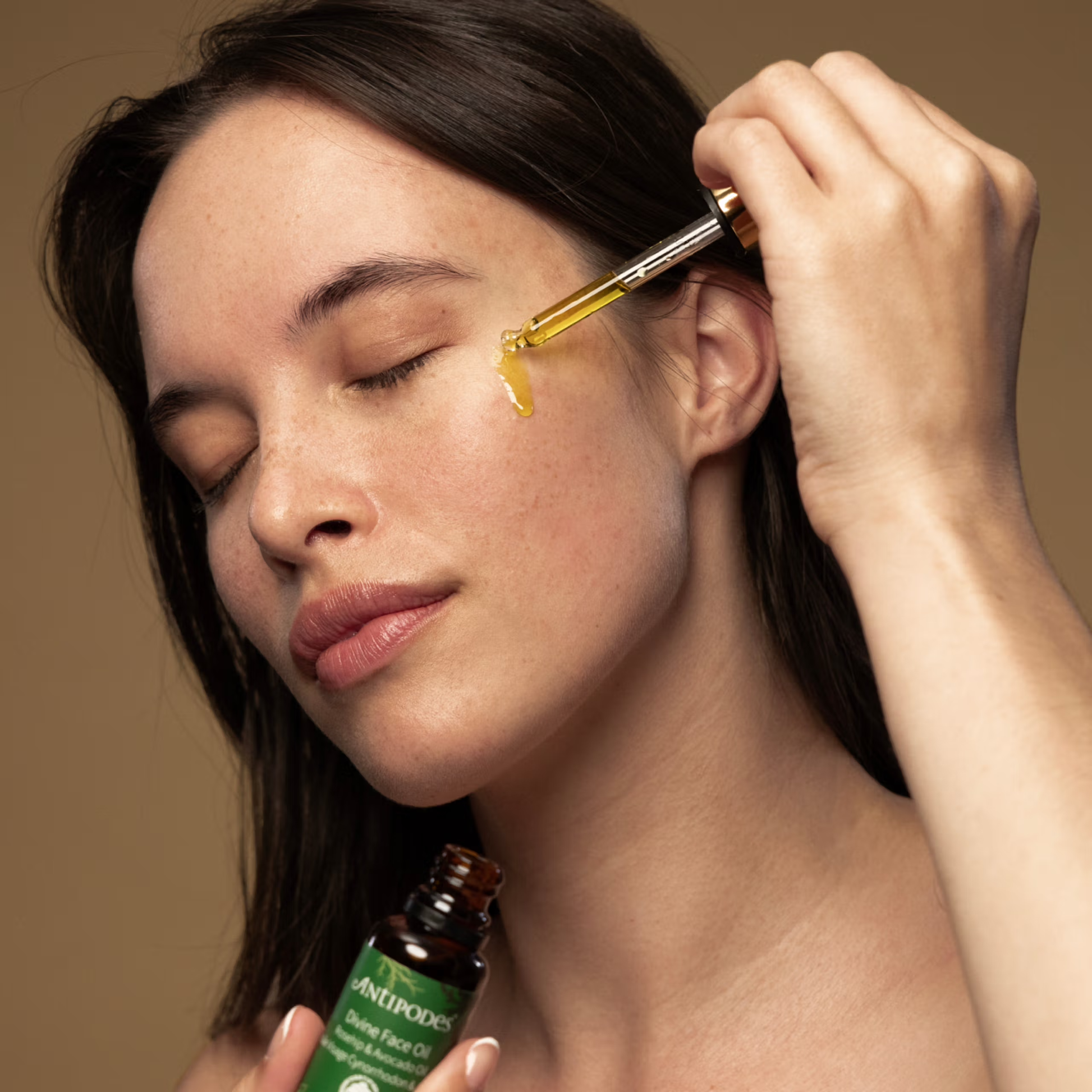 Woman applying a dropper of oil to her face with a bottle labeled 'Anthropologie' in the foreground.
