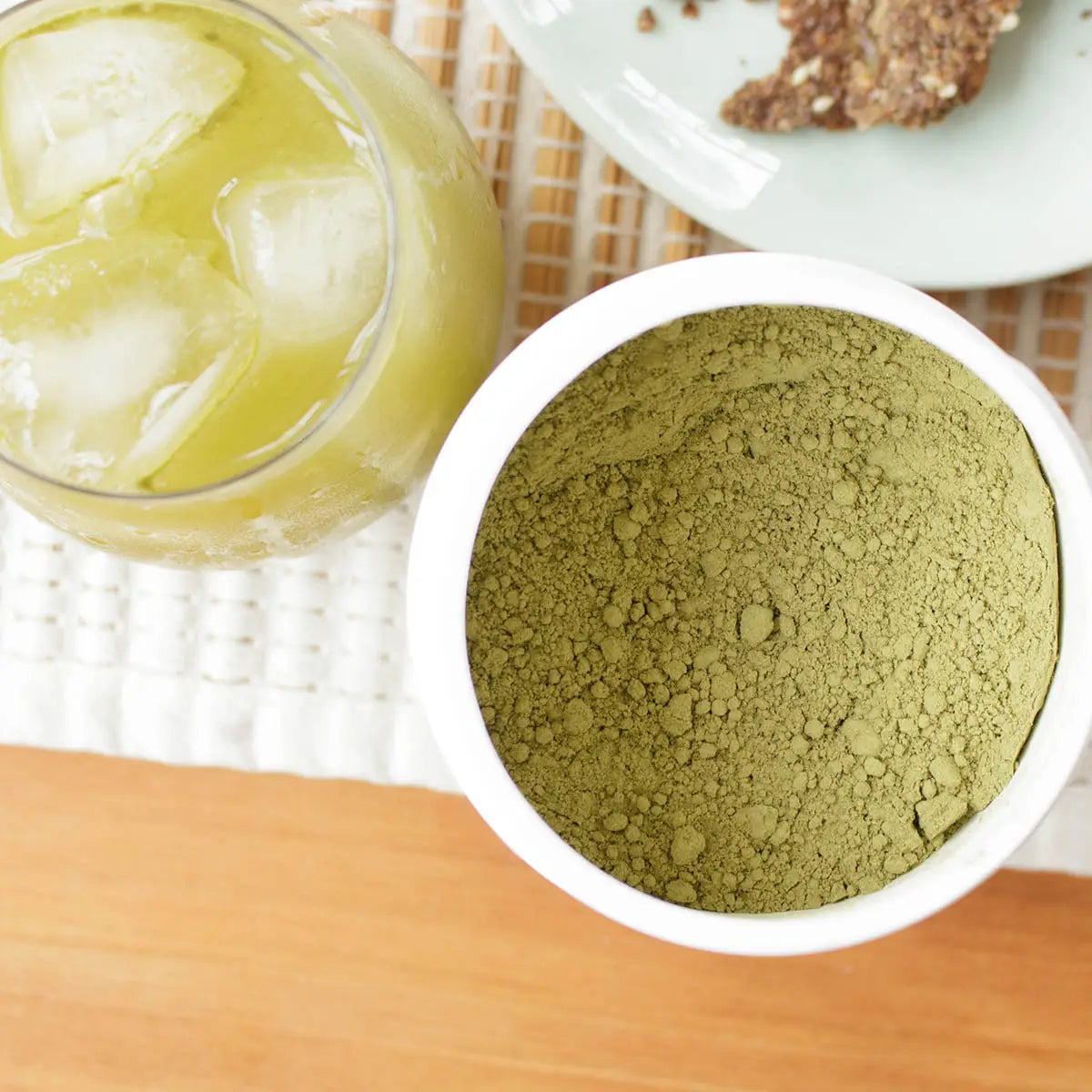 Moringa powder in a white container with a glass of iced moringa drink on a wooden surface.