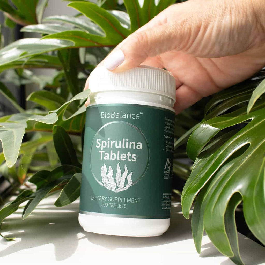 BioBalance Spirulina 500 Tablets bottle held by a hand with green leaves in the background.