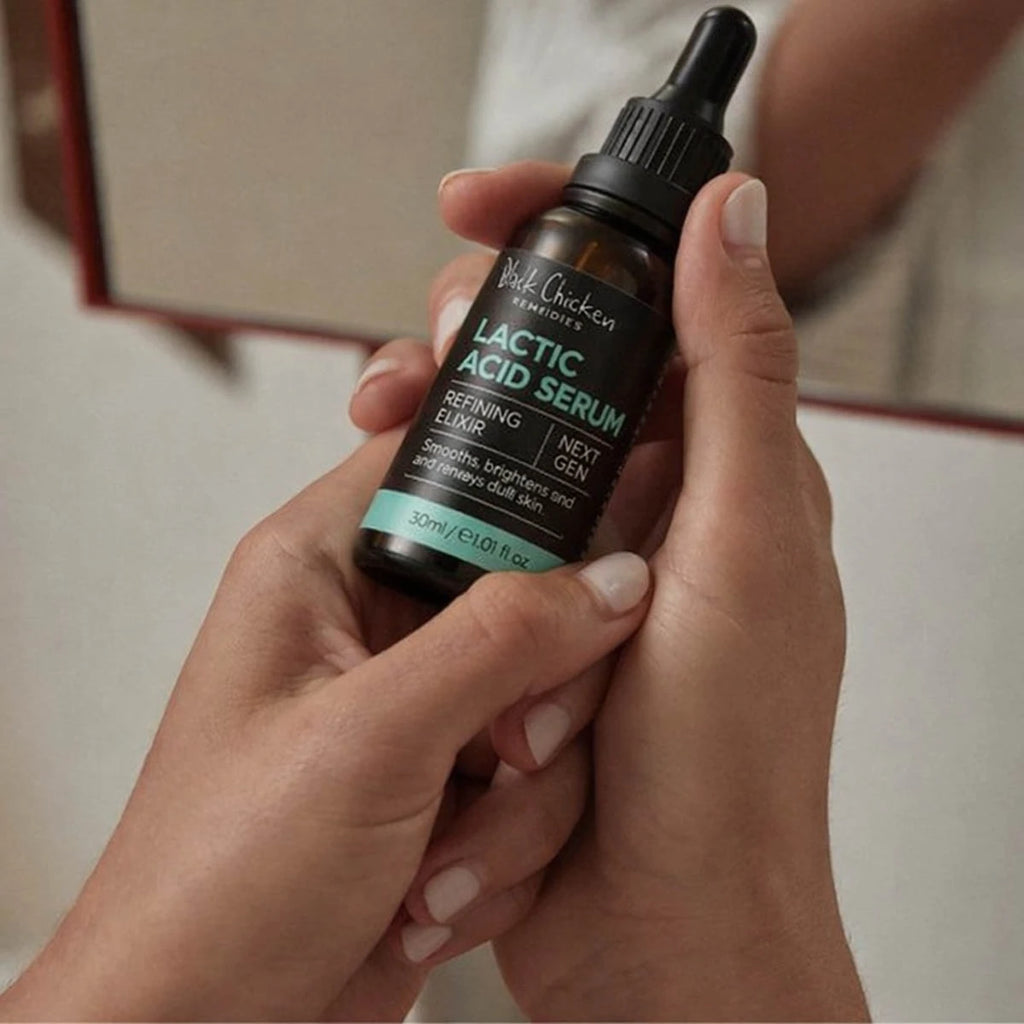 A person holding a small amber dropper bottle labeled 'Black Chicken Remedies Lactic Acid Serum' against a softly blurred indoor background.