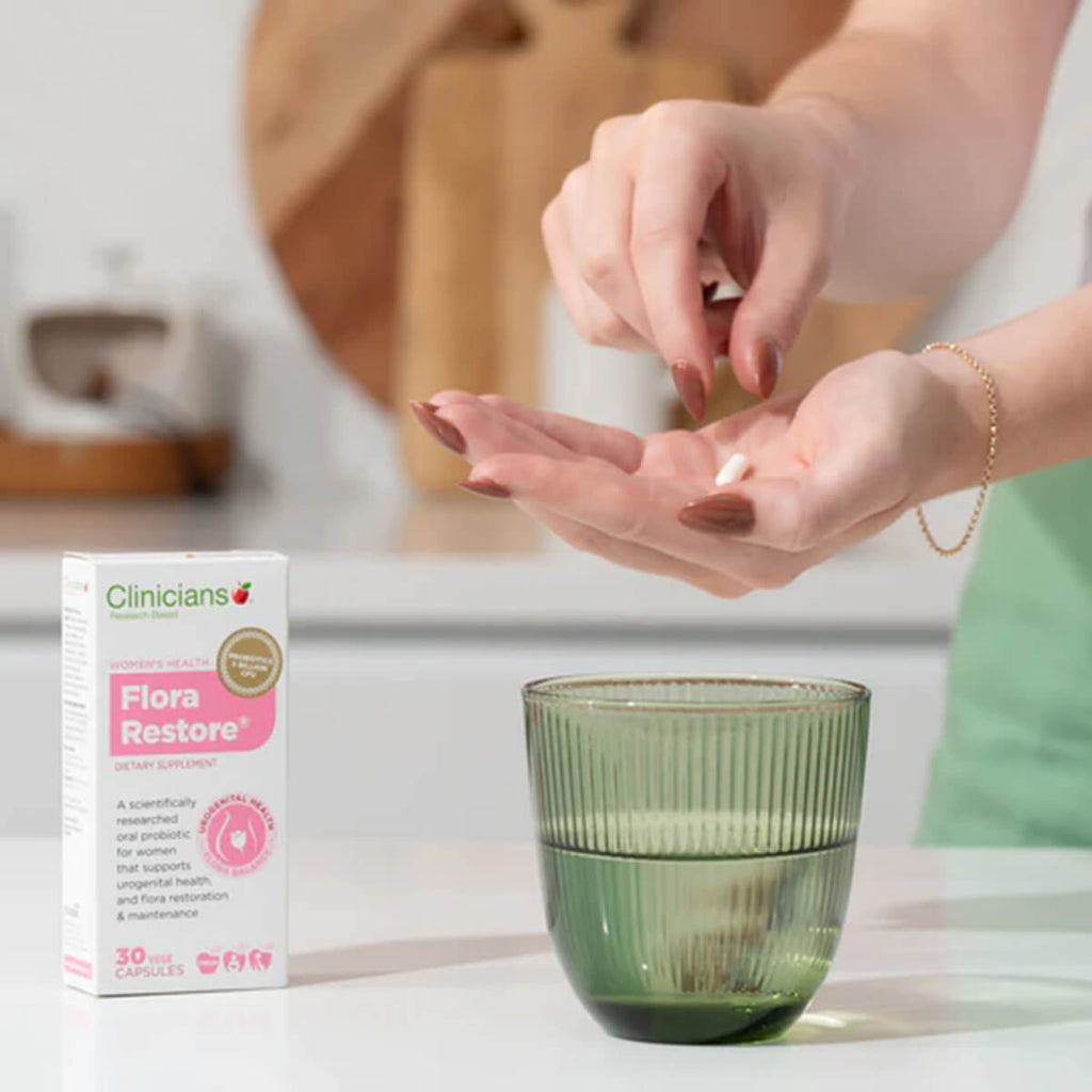 Person holding a pill with Clinicians Flora Restore packaging and a green glass in the background.