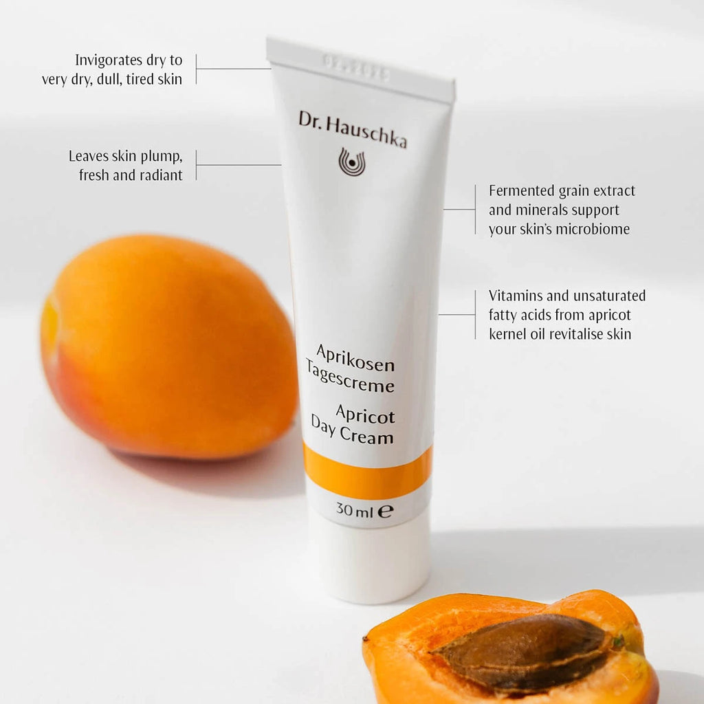 A white tube of Dr. Hauschka Apricot Day Cream stands on a white surface with whole and halved apricots in the background and callout text describing benefits.