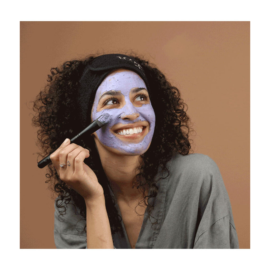Woman applying a purple facial mask with a brush on a brown background.