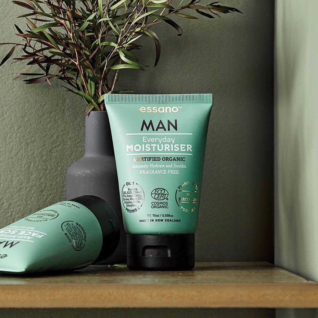 Green tube of essano Man Everyday Moisturiser on a wooden surface with a vase and plant in the background.
