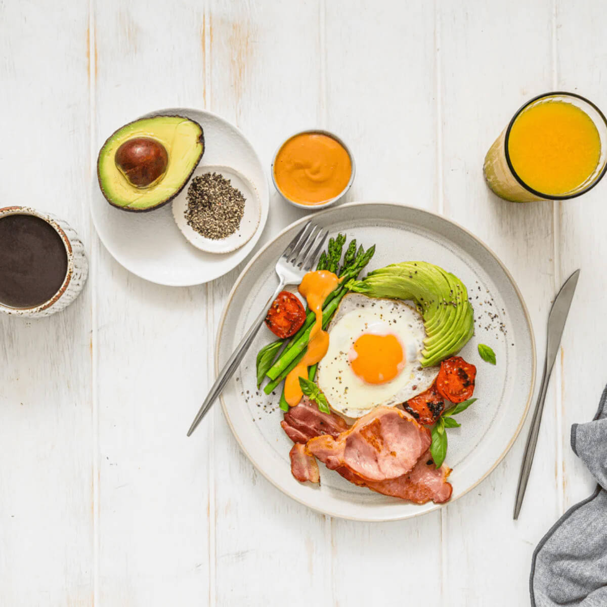 Breakfast plate with eggs, bacon, avocado, and vegetables on a white wooden table.