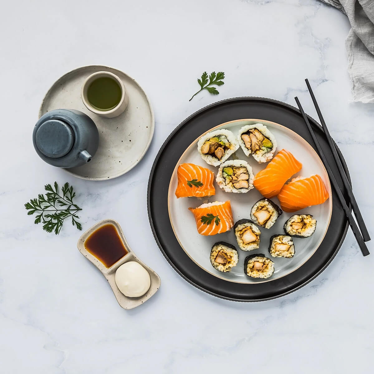 Assorted sushi on a plate with a cup of green tea, soy sauce, and wasabi on a light gray surface.