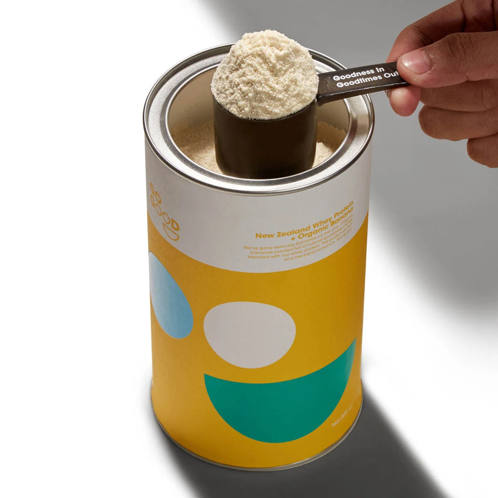 Canister of banana protein powder with a scoop being taken out, on a neutral background