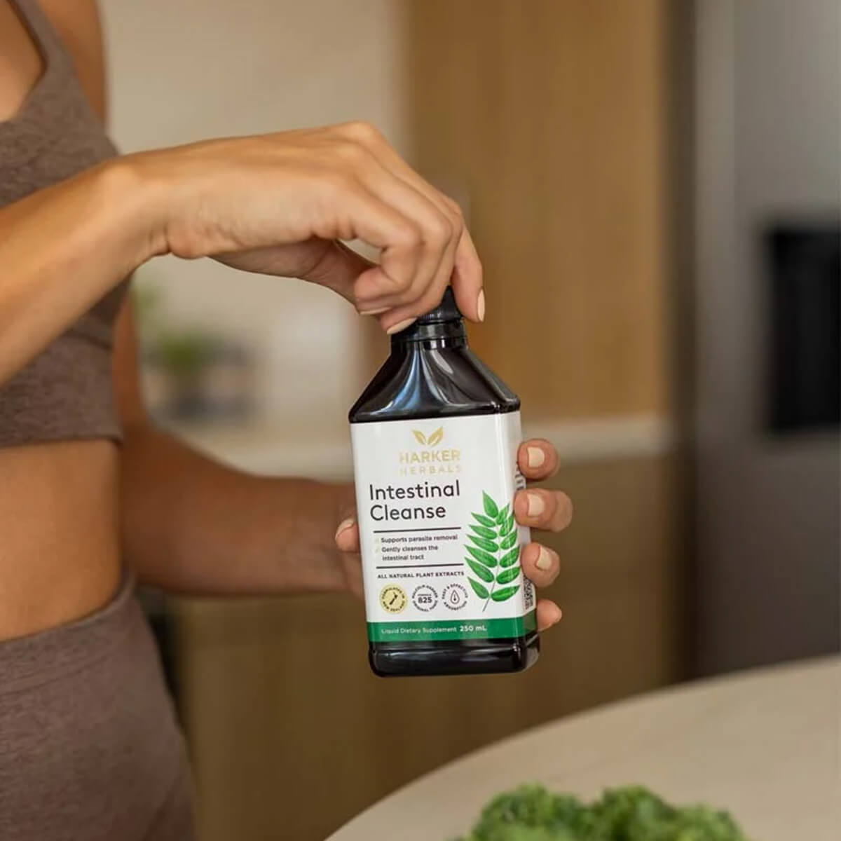 A person in a brown sports bra holds a bottle of Intestinal Cleanse in a kitchen setting.