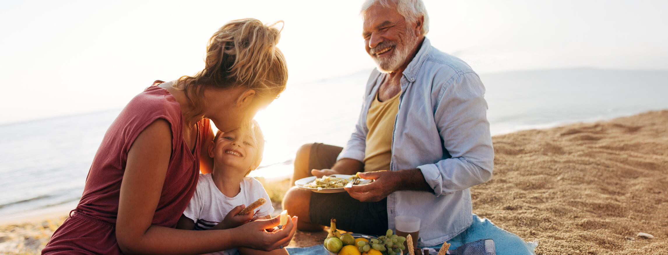 Man and two children enjoying a picnic on a sandy beach with ocean view.